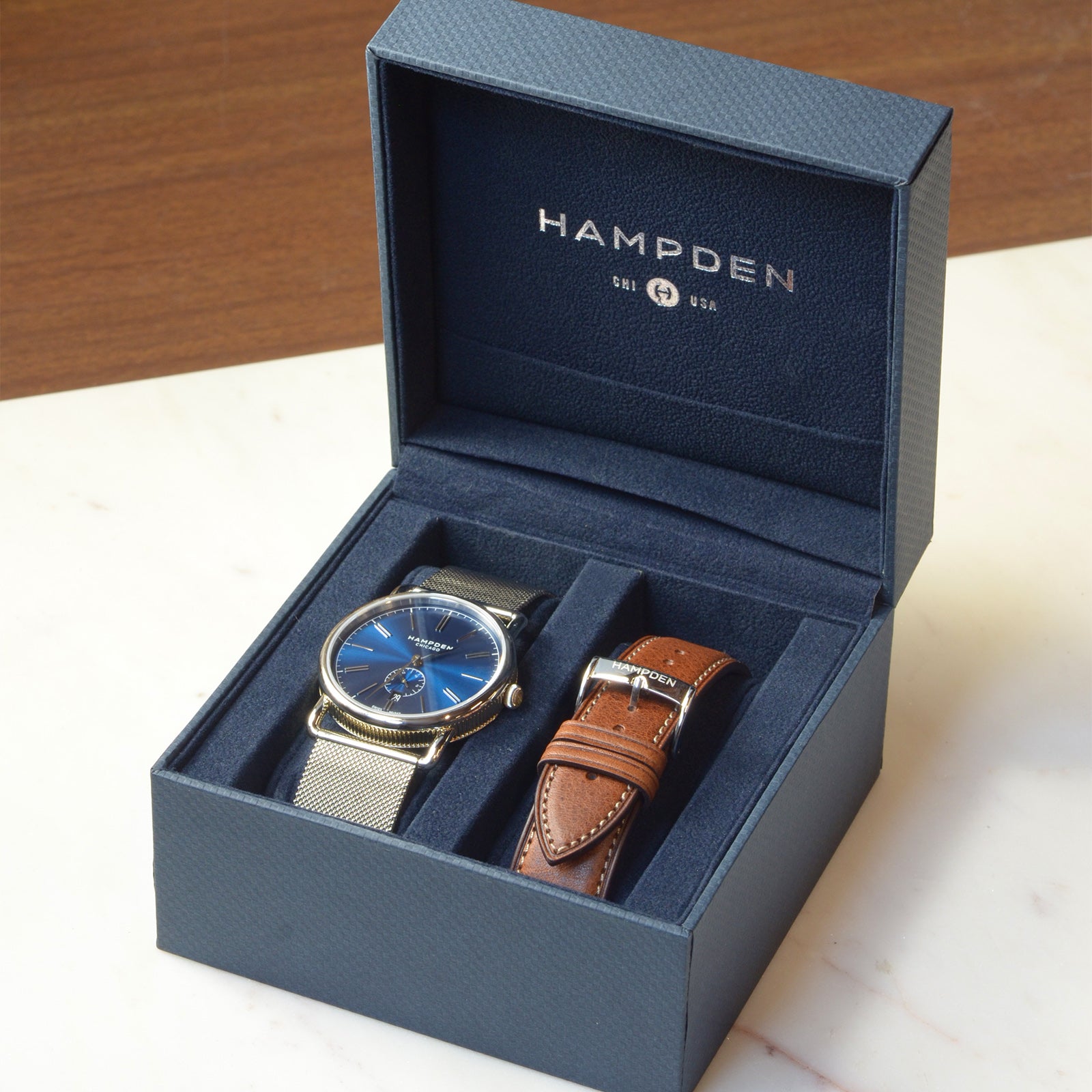 An open navy Hampden presentation box on a table, holding a blue dial watch and a spare tan leather strap.